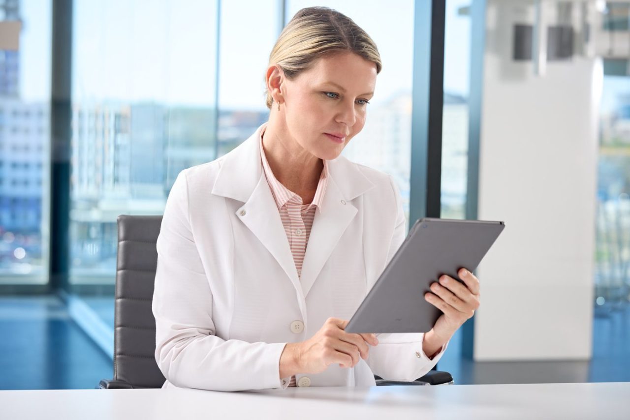 A person wearing a white coat sits at a desk, holding and looking at a tablet. The background features large windows with a view of buildings. The individual appears focused and professional.