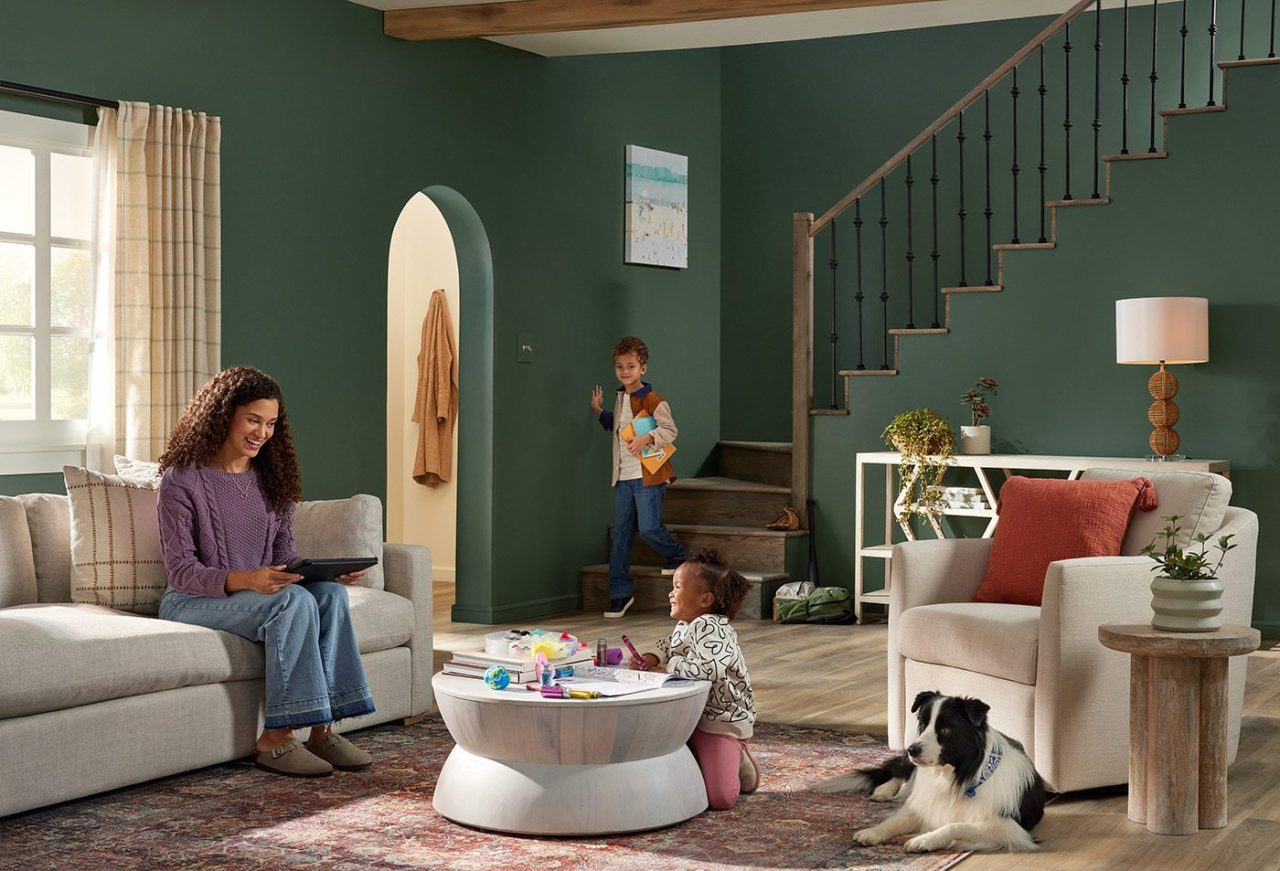 A cozy living room boasts green walls and a staircase, reflecting thoughtful set design. A woman sits on a sofa engrossed in her tablet, while a child plays with toys on the floor and another stands on the stairs. Nearby, a black-and-white dog rests amidst plants and decorative items.