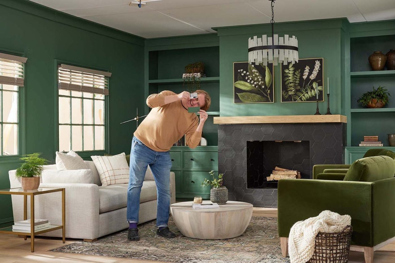 A person takes a photo with a smartphone, leaning creatively amidst a set design in the living room. The space features green walls, a white sofa, green armchair, and a round wooden coffee table. The fireplace showcases hexagonal tiles topped with nature-themed artwork.
