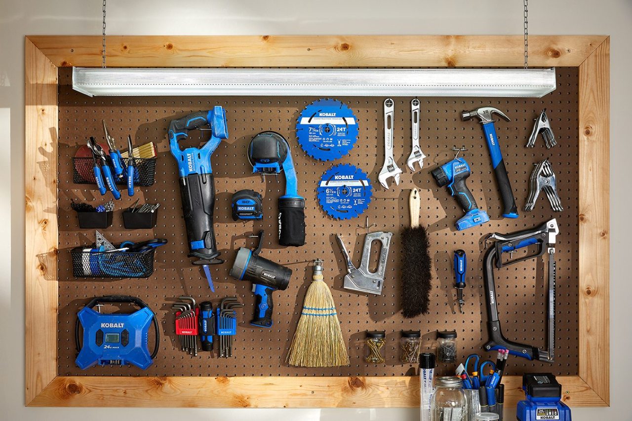 A wall-mounted wooden pegboard with various hand tools, including a drill, wrench, screwdriver set, and circular saw blades. The tools are organized neatly, with a broom and other accessories hanging beside them.