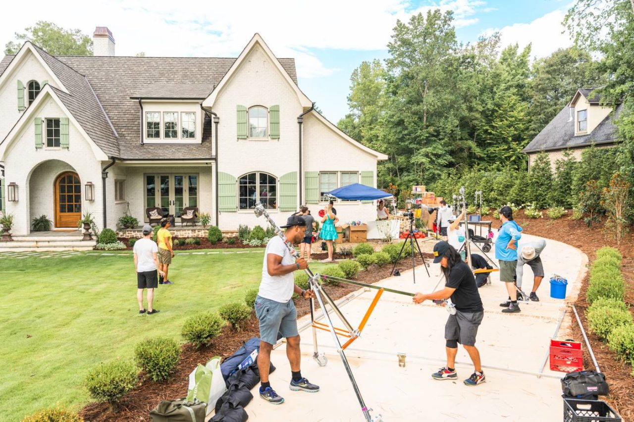 A film crew is setting up equipment in the yard of a large, light-colored house with green shutters. Several people are arranging cameras and other gear, while others stand nearby. The scene is lively and surrounded by greenery.