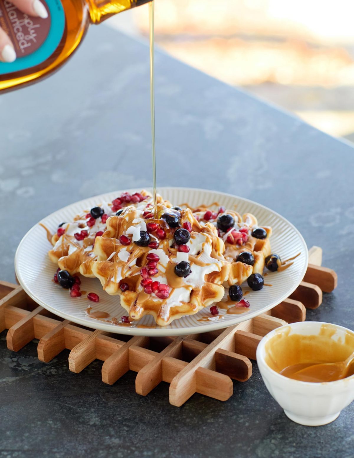 a plate of waffles topped with blueberries and pomegranate seeds being drizzled with syrup