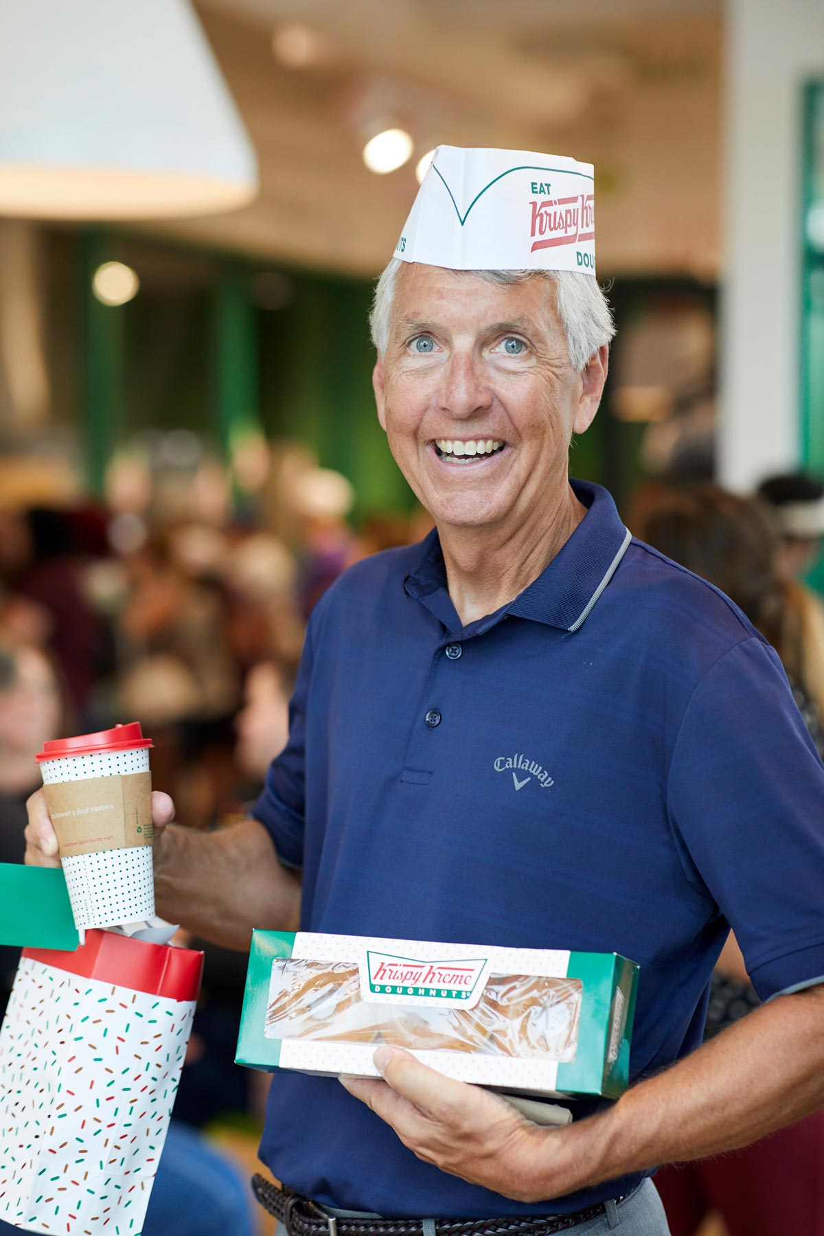 Older man with white hair wearing a Krispy Kreme paper had and holding a package of doughnuts and a coffee
