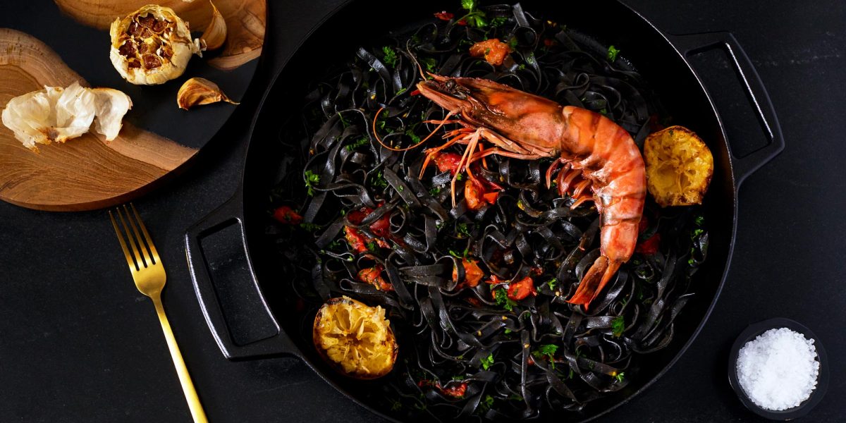 Dramatic lifestyle food photography of a bright red African Prawn placed on top of black squid ink pasta garnished with roasted garlic
